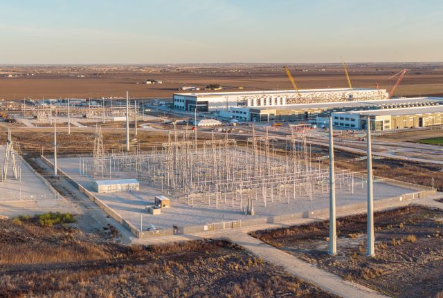 An aerial view of a power substation with tall structures and a large industrial building in the background.