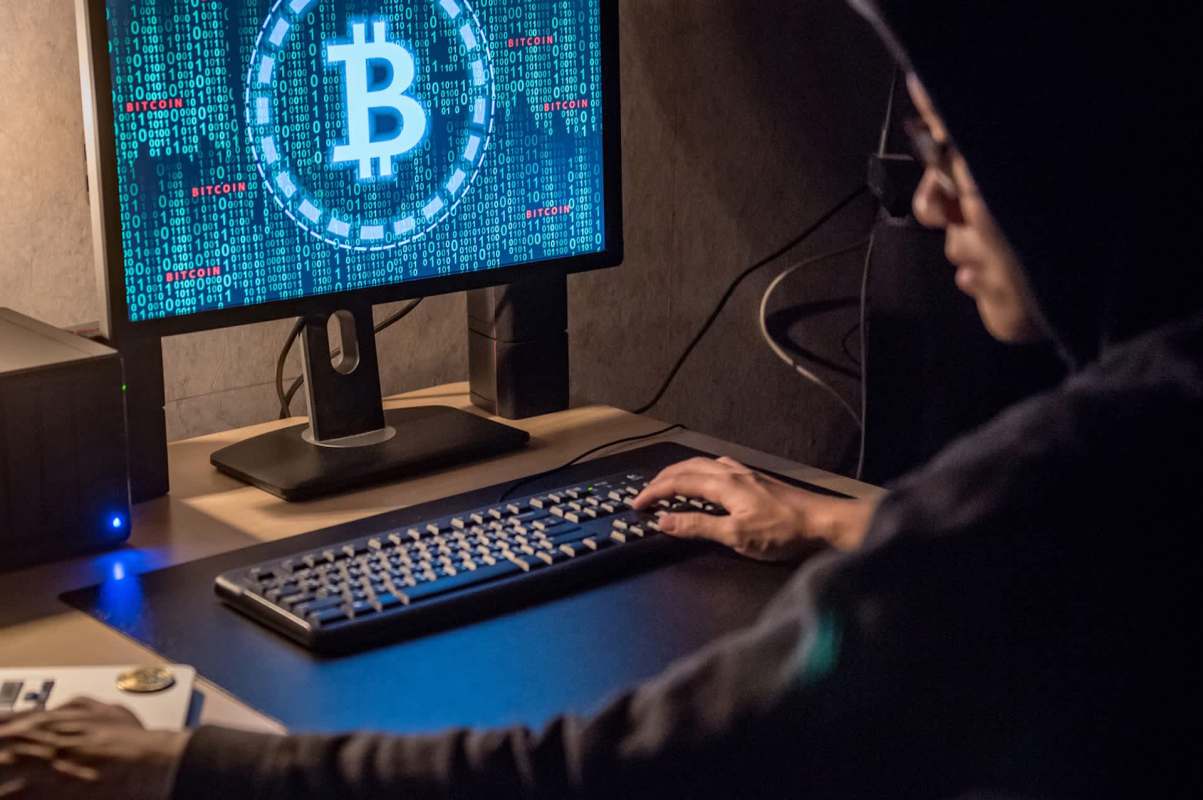A person in a hoodie uses a computer to access cryptocurrency data on a dark, illuminated workspace.