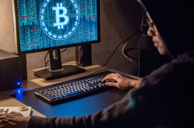 A person in a hoodie uses a computer to access cryptocurrency data on a dark, illuminated workspace.