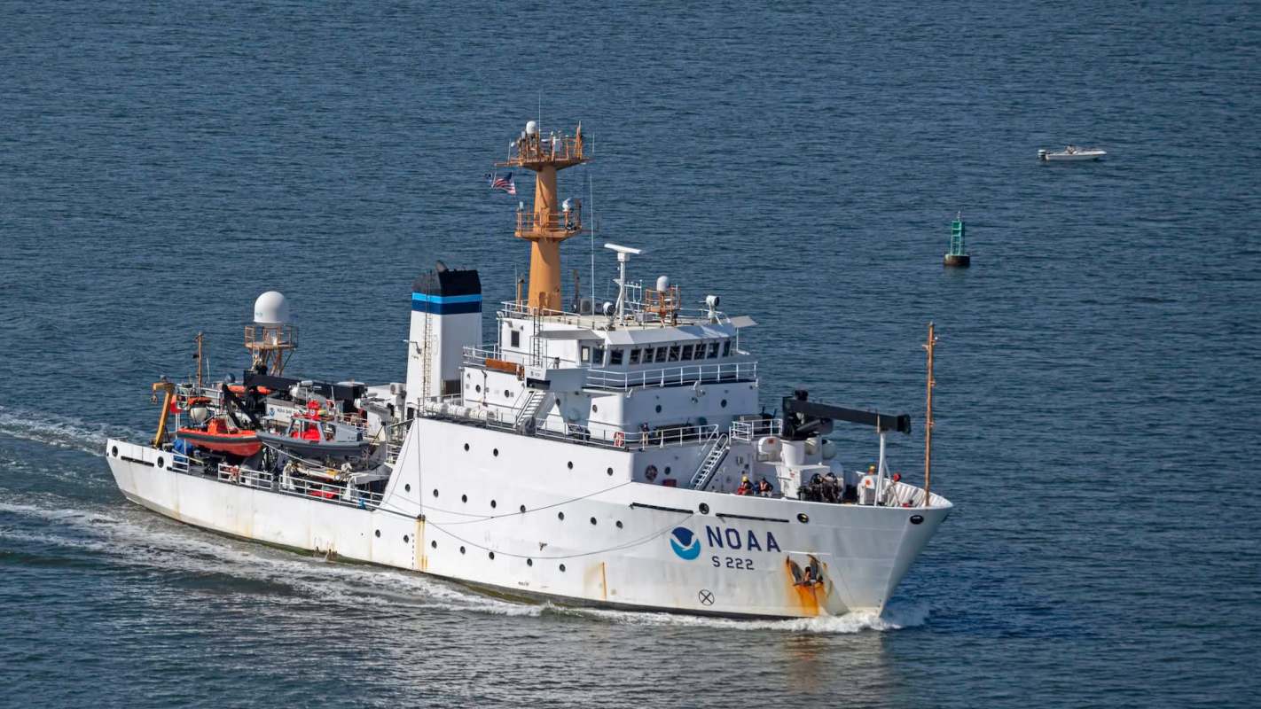An NOAA research ship navigates through calm waters with small boats and a buoy nearby.