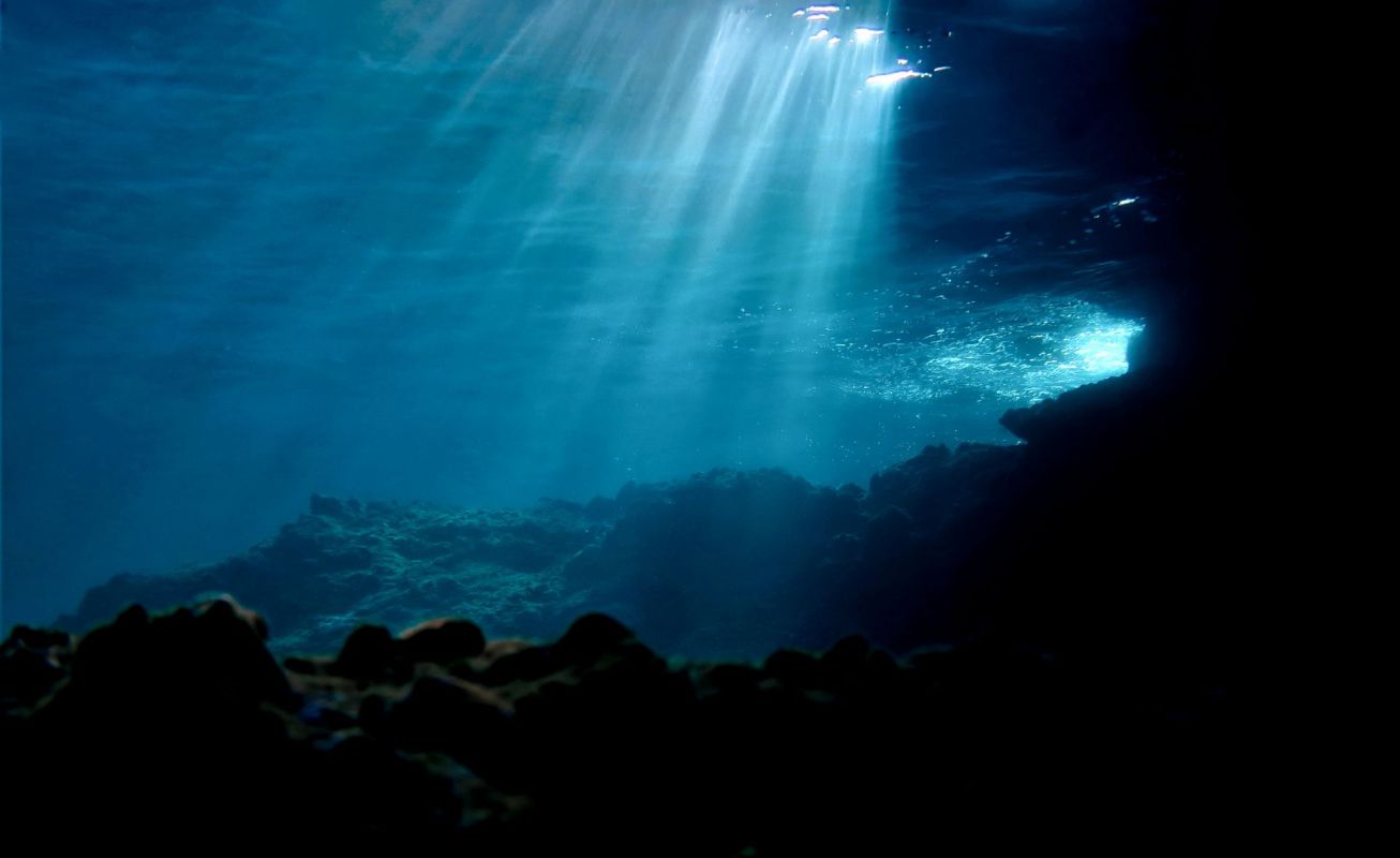 Ray of sunlight streaming through the water into a deep blue underwater scene with rocky formations.
