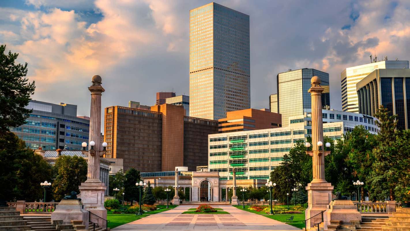 A city park lined with columns and greenery, framed by modern skyscrapers under a partly cloudy sky.