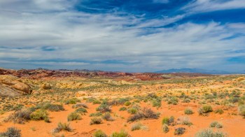 A vast desert landscape featuring colorful rock formations under a cloudy blue sky.