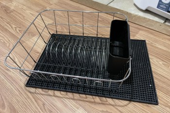 A metal dish rack on a black drying mat, placed on a wooden floor.