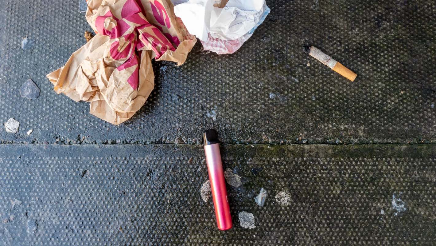 A pink vape pen lies on a black surface beside trash.