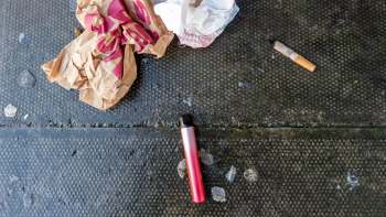 A pink vape pen lies on a black surface beside trash.