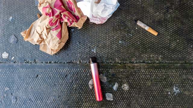 A pink vape pen lies on a black surface beside trash.