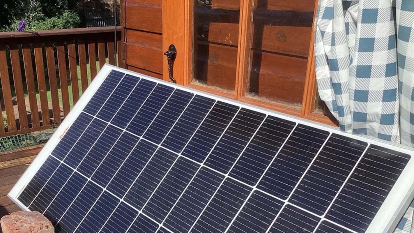 A solar panel is propped against a wooden wall near a window with checked curtains.