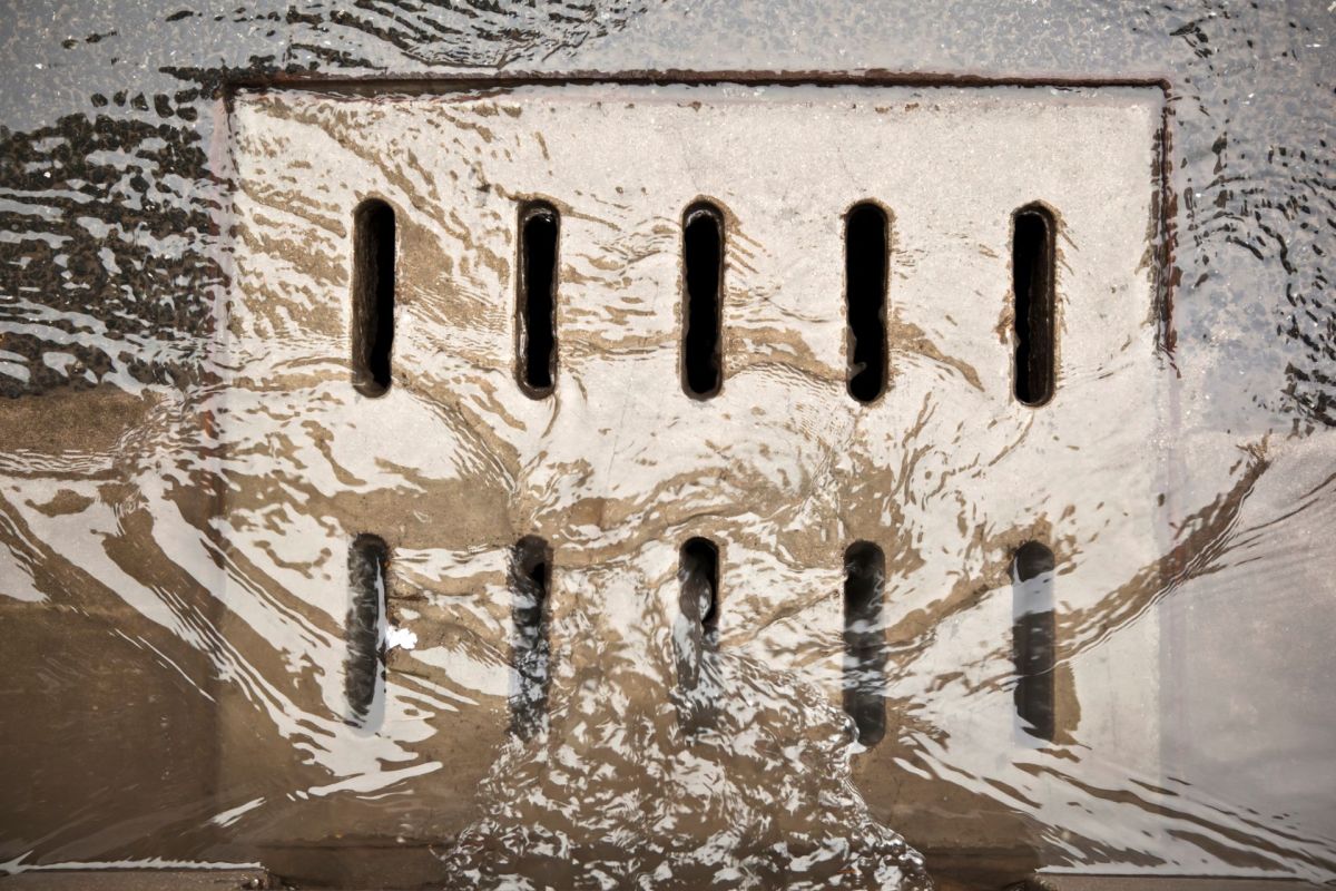 A close-up of a drainage grate partially submerged in flowing water with ripples around it.