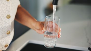 A person is standing at a sink, filling a glass with tap water.