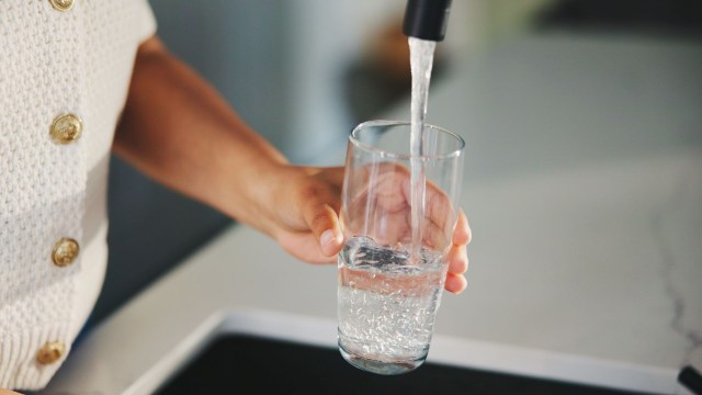 A person is standing at a sink, filling a glass with tap water.