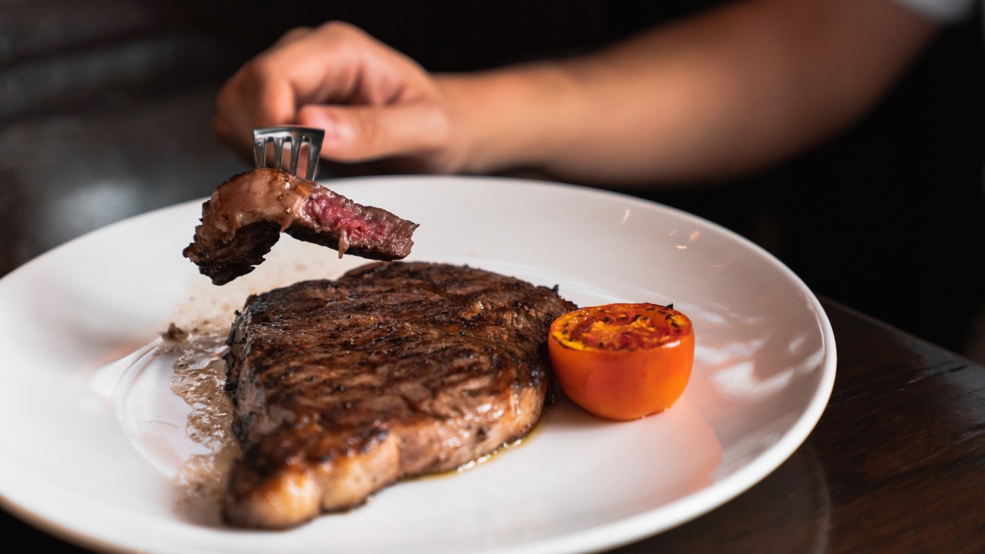 A fork lifts a bite of steak from a plate with steak and grilled tomato.