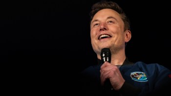 SpaceX CEO Elon Musk is smiling and speaking into a microphone while wearing a dark jacket with a patch.