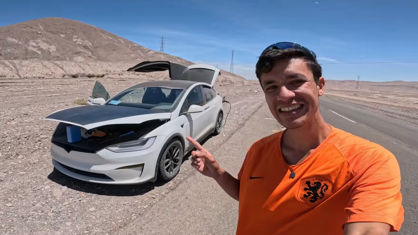 A smiling man points at a white Tesla Model X with its front trunk open, set against a rocky landscape.