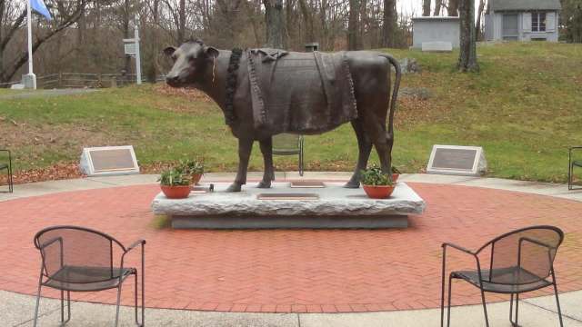 A bronze statue of a cow stands on a stone pedestal in a circular brick plaza surrounded by potted plants.
