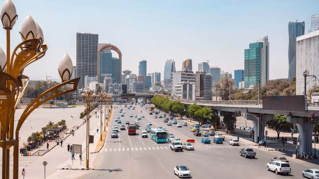 A busy urban street featuring traffic, modern buildings, and decorative streetlights on a sunny day.