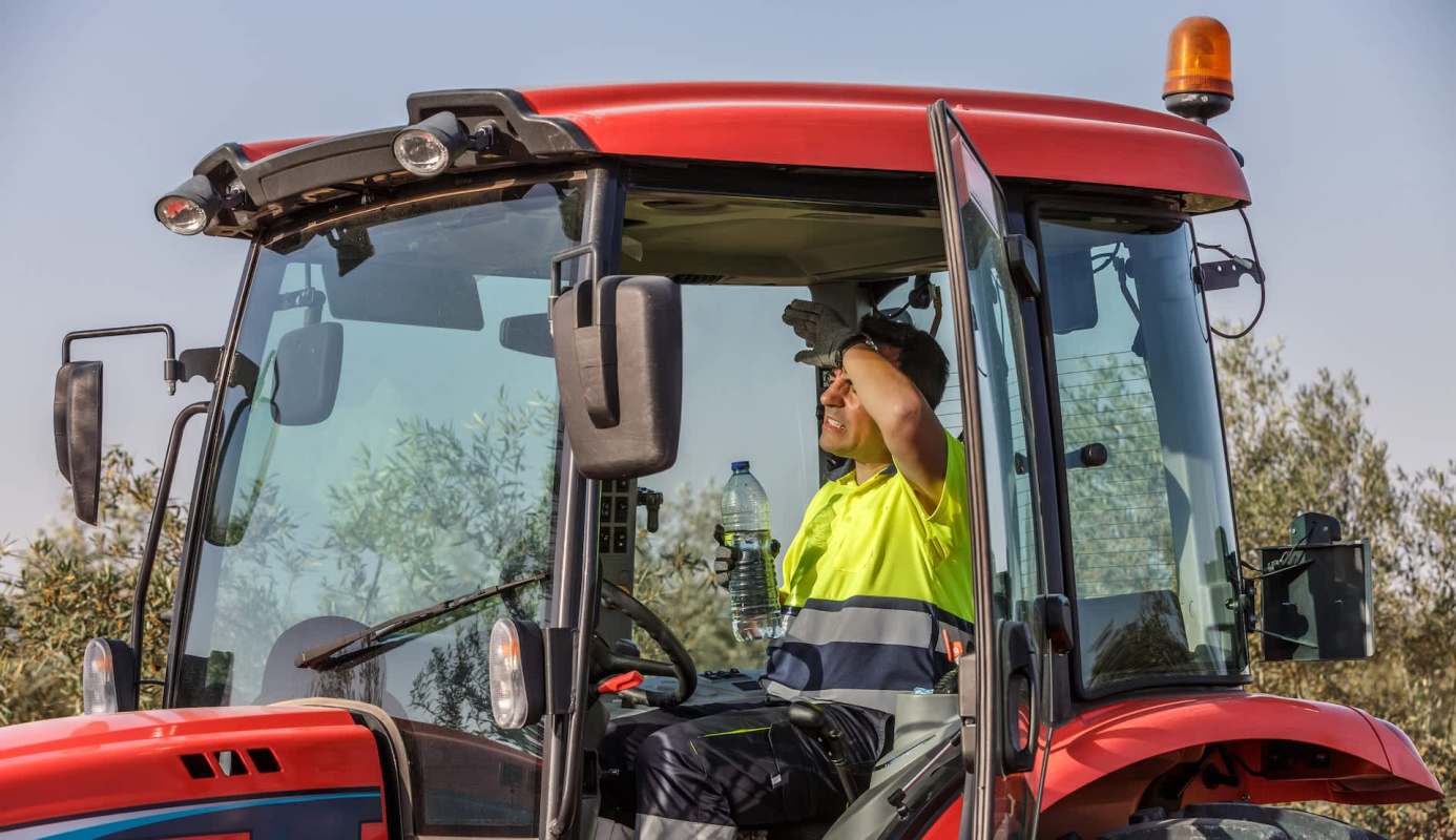 A man in a bright yellow shirt sits in a red tractor, wiping sweat from his forehead while holding a water bottle.