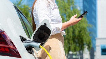 A woman holding a smartphone stands beside an electric vehicle connected to a charging station.