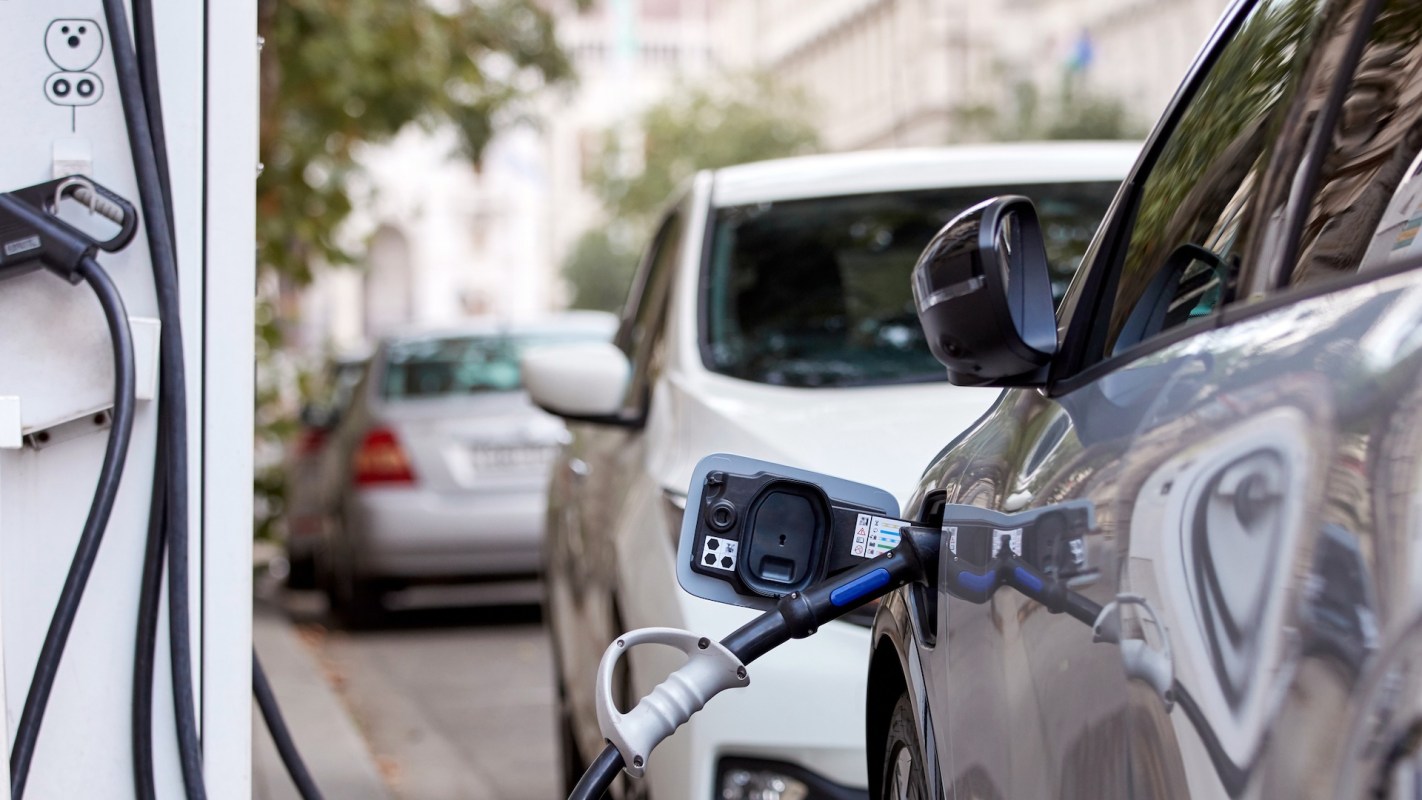 An electric vehicle is plugged into a charging station on a city street with parked cars in the background.