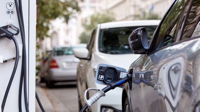 An electric vehicle is plugged into a charging station on a city street with parked cars in the background.