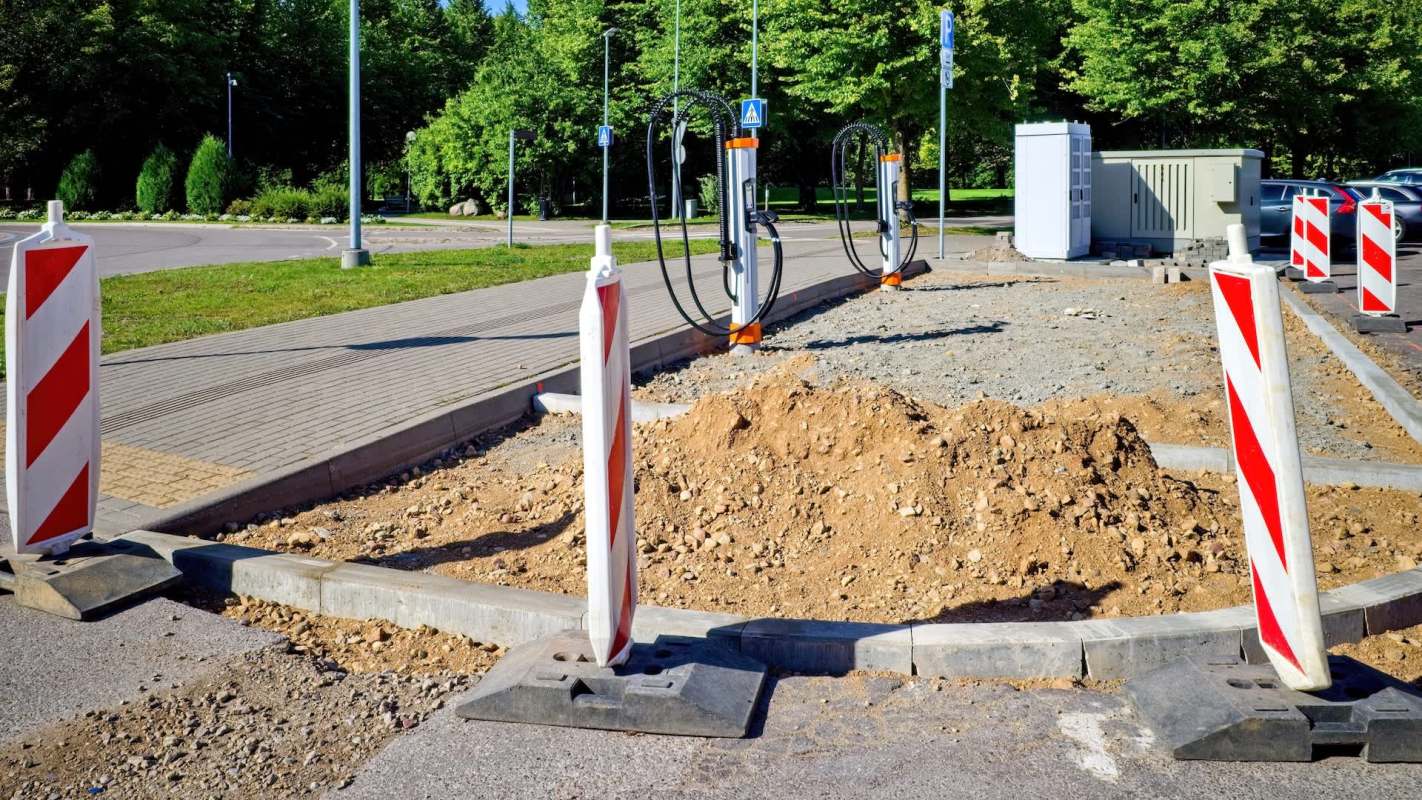 Construction site with orange charging stations, dirt piles, and warning barriers on a paved area surrounded by greenery.