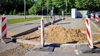 Construction site with orange charging stations, dirt piles, and warning barriers on a paved area surrounded by greenery.