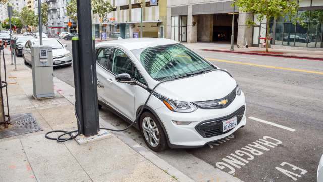 A white electric vehicle is charging at a street charging station in an urban setting.