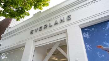 A storefront of Everlane with a white facade and large window, partially obscured by tree branches.