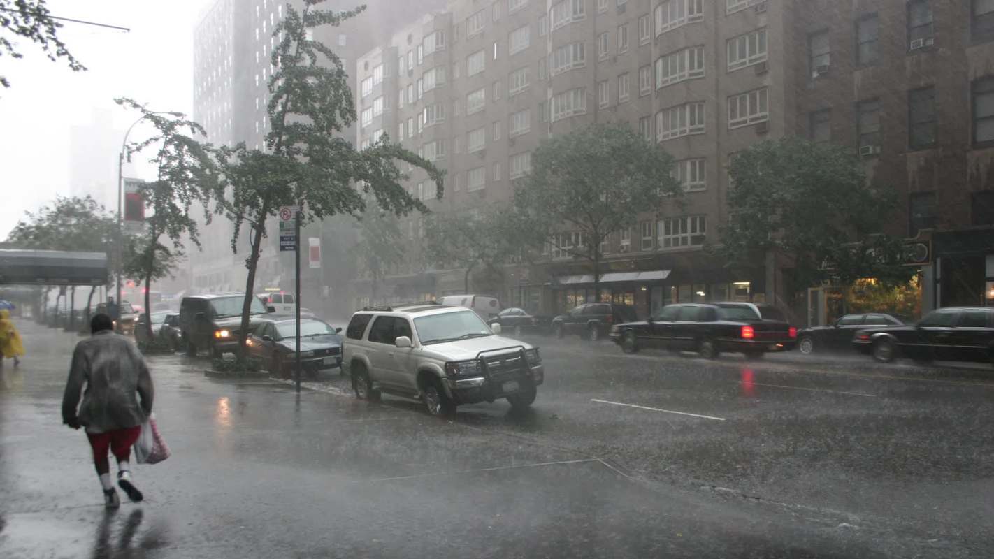 Heavy rainfall creates a wet street scene as a person hurries by, with cars and buildings in the background.