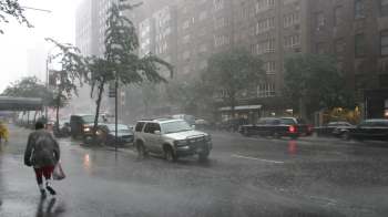 Heavy rainfall creates a wet street scene as a person hurries by, with cars and buildings in the background.