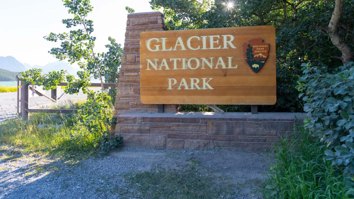 A wooden sign for Glacier National Park surrounded by greenery and mountains in the background.