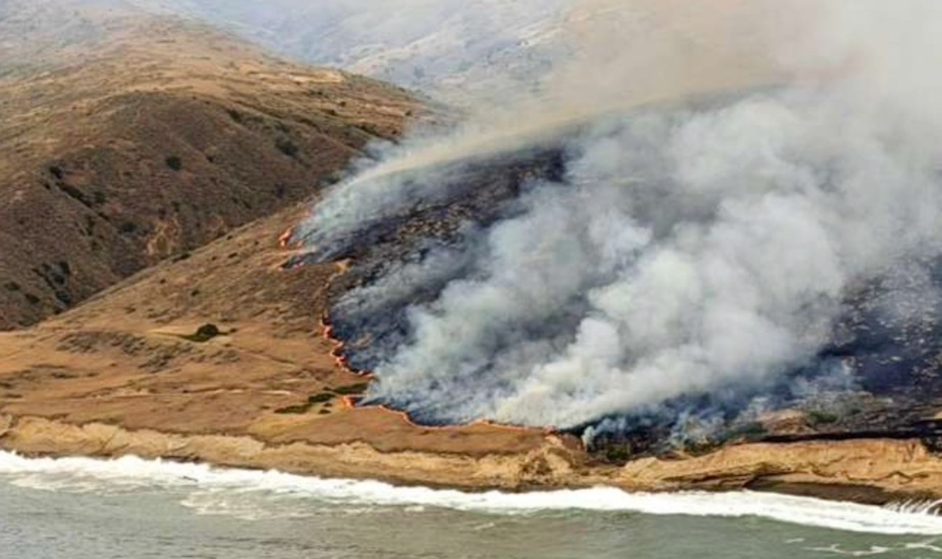 A hillside on fire with smoke billowing over a coastal landscape and crashing waves below.