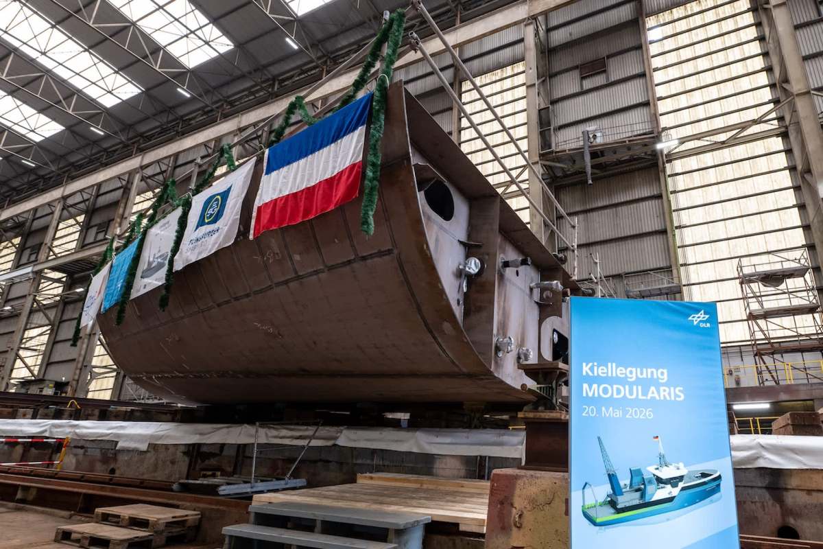 A large ship hull is displayed in a factory, adorned with flags and a sign announcing its launch date in 2026.