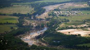 An aerial view of a river winding through a landscape featuring greenery, dry areas, and nearby housing.