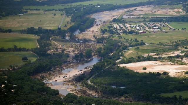 An aerial view of a river winding through a landscape featuring greenery, dry areas, and nearby housing.