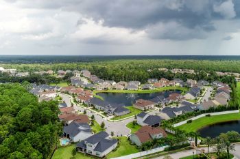 An aerial view of a suburban neighborhood with houses, streets, and a central pond surrounded by trees.