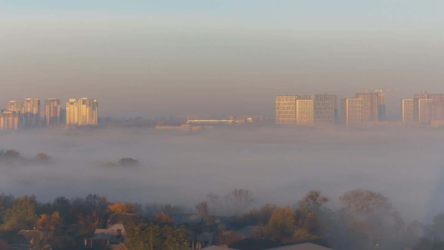 A city skyline partially obscured by morning fog, with the sun illuminating the buildings.
