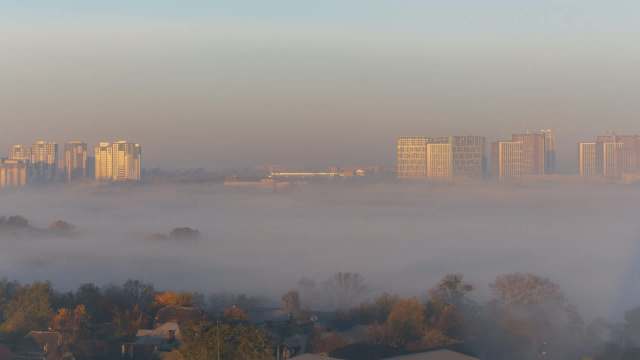 A city skyline partially obscured by morning fog, with the sun illuminating the buildings.