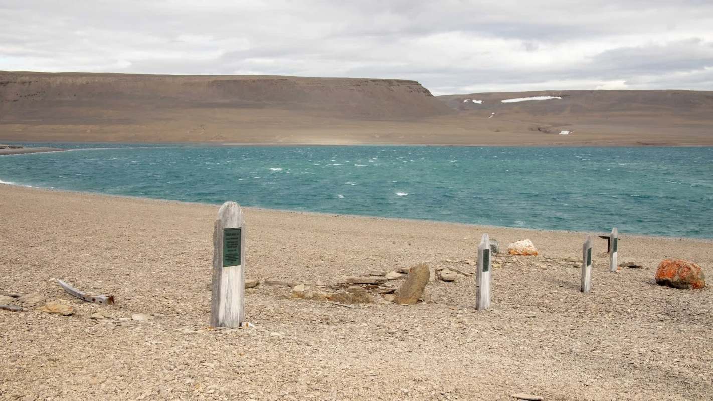A stony beach with wooden headstones and a turquoise ocean in the background under a cloudy sky.
