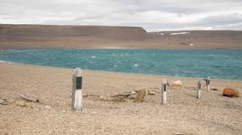 A stony beach with wooden headstones and a turquoise ocean in the background under a cloudy sky.
