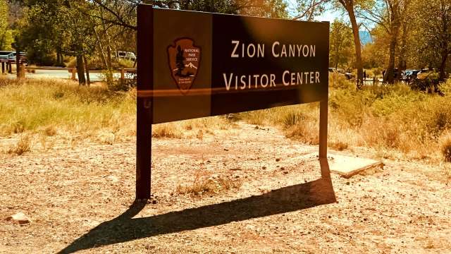 A sign for the Zion Canyon Visitor Center surrounded by grass and trees in a sunny landscape.