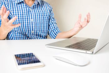 A man in a checkered shirt gestures with frustration while sitting at a desk with a laptop and smartphone.