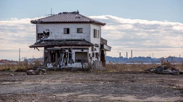 An abandoned and dilapidated two-story house stands in a barren landscape under a cloudy sky.