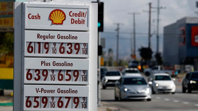 A gas station sign displays fuel prices for regular, plus, and V-Power gasoline with cars driving in the background.