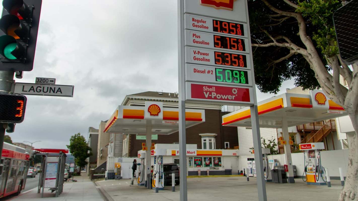 A Shell gas station displays fuel prices and a traffic light at an intersection.