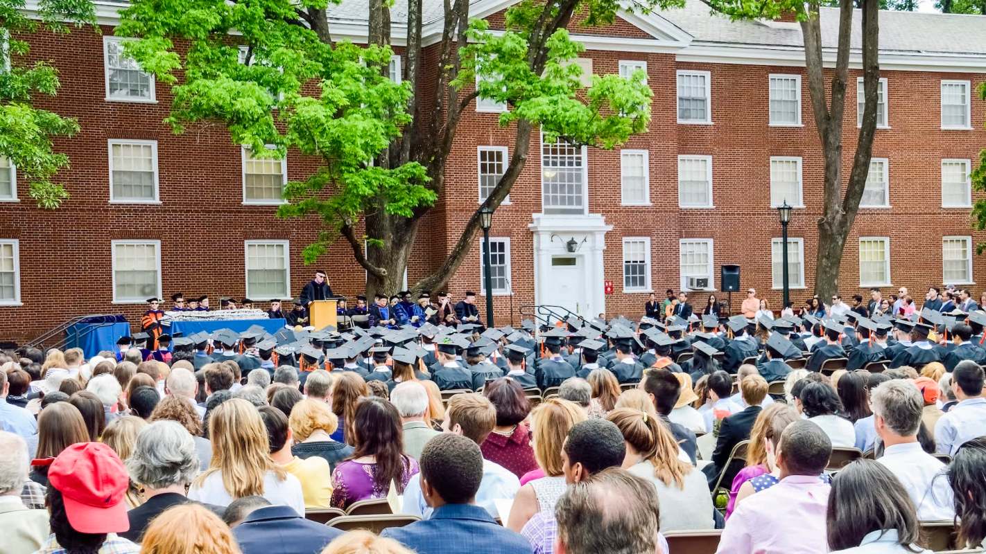 A large crowd gathers for a graduation ceremony outdoors, with graduates in caps facing a speaker on a podium.