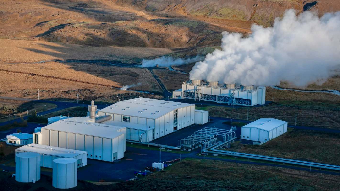 A geothermal power plant with steam rising, surrounded by rocky terrain and grassy land.