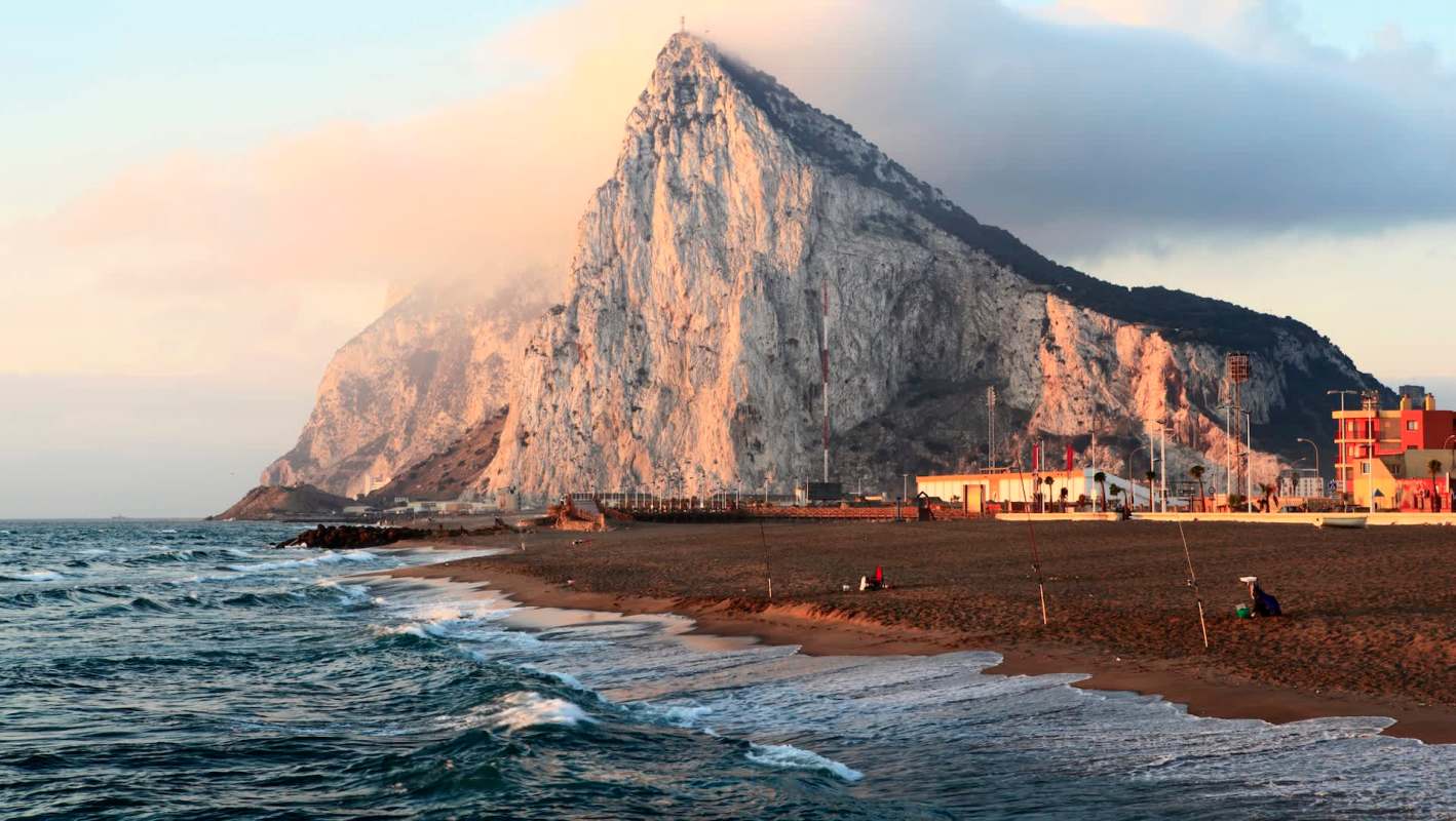 A sandy beach with gentle waves, the towering rock of Gibraltar in the background, and a few people fishing along the shore.