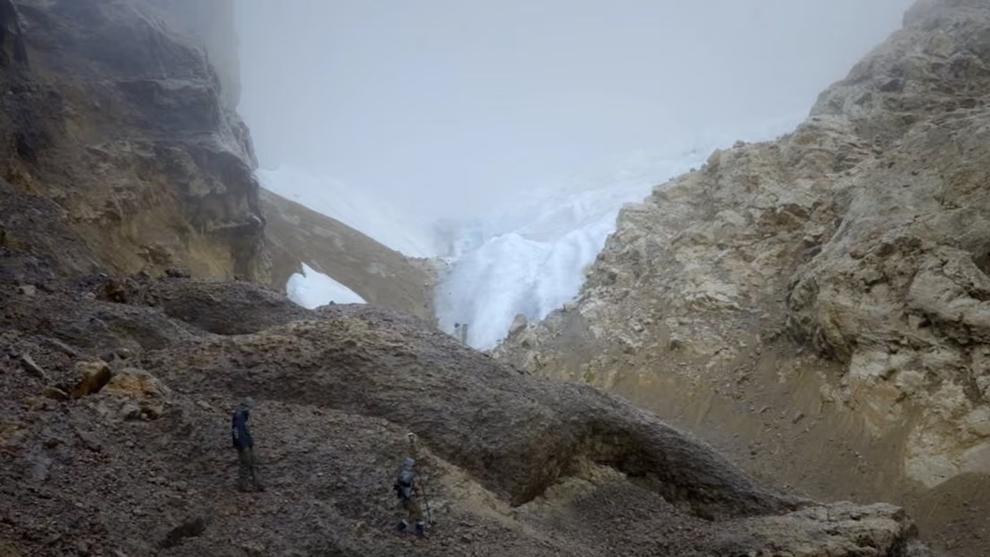 A rocky valley with two climbers near a glacier, shrouded in fog and surrounded by steep cliffs.