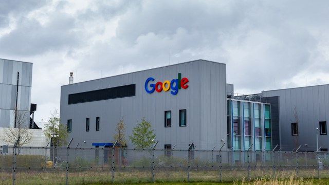 A Google-branded building is shown with a gray exterior and colorful logo under a cloudy sky.